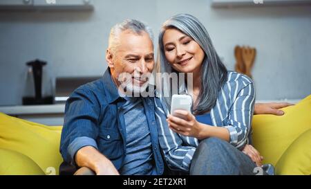 Lächelnde asiatische grauhaarige Frau mit Smartphone in der Nähe glücklich Ehemann Stockfoto