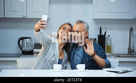 Fröhlicher älterer Mann winkt Hand in der Nähe asiatische Frau, die Selfie In der Küche Stockfoto