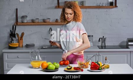 Lockige Frau hält Notizbuch und wiegt Tomaten in der Nähe von Gemüse Und Orangensaft auf dem Tisch Stockfoto