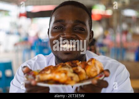 Afrikanischer Mann, der ein gegrilltes Hähnchen auf dem Gericht mit aufhebt Lächeln und happy,16:9 Stil Stockfoto