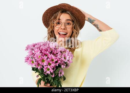 Fröhliche junge Frau im Sommer Strohhut und Brillen halten Bouquet von rosa Blumen stehen auf weißem Hintergrund. Stockfoto