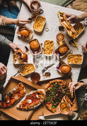 Lockdown Familie Fast Food Abendessen von Lieferservice. Flat-Lay von Freunden oder Verwandten Hände mit Quarantäne-Home-Party mit Burger, Pommes, Sandwiches, Pizza, Bier über Tisch Hintergrund, Draufsicht Stockfoto