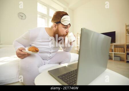 Lustige bärtige übergewichtige männliche sitzen mit Kaffee und Brötchen für Breakfasy und Film auf dem Laptop Stockfoto