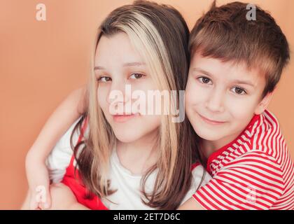 Porträt von Kindern auf braunem Hintergrund. Der jüngere Bruder umarmt seine ältere Schwester mit Zärtlichkeit und Liebe Stockfoto