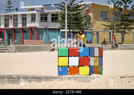 MAROUBRA, AUSTRALIEN - 04. Dez 2015: Professioneller Fotograf sitzt auf der ikonischen Rubik's Cube Skulptur am australischen Strand von Maroubra Stockfoto