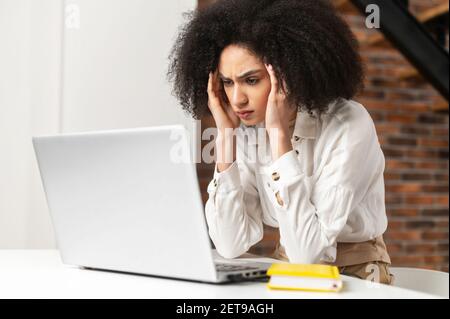 Desorientierte junge Frau im Büro unter emotionalem Stress, Blick auf Laptop, ihre Hände an seinen Schläfen halten, Falten die Stirn, überarbeitet, Computer ist gebrochen, was passiert ist Stockfoto