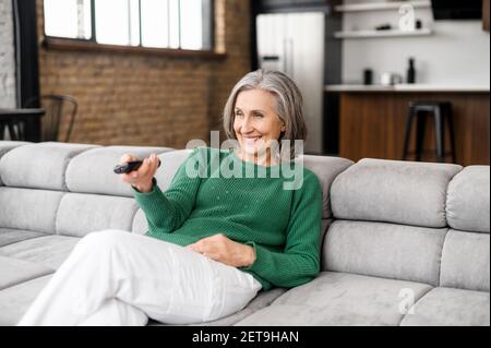 Glückliche lächelnde ältere Dame mit grauen Haaren in einem grünen Pullover sitzt auf der Couch im Wohnzimmer, zeigt eine tv-Fernbedienung, um die Kanäle zu wechseln, aufgeregt, um Lieblings-Film Online-Serie zu sehen Stockfoto