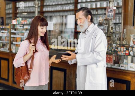 Erfahrene professionelle bärtige ältere Mann Apotheker hält Tablette und berät junge Dame Kunden in alten alten alten Drogerie. Apotheker und Patient Stockfoto