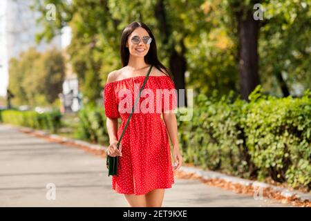 Foto Porträt von weiblichen Studenten lächelnd tragen stilvolle rote Kleid Im Sommer heiße Wetter Sonnenbrille Wandern im grünen Park Stockfoto