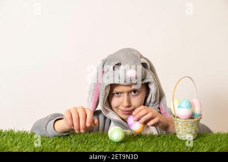 Ein Mädchen in einem Kaninchenkostüm sieht sich Ostereier auf dem grünen Gras an und ist überrascht. Isoliert auf Weiß. Osterfeiertagskonzept. Stockfoto