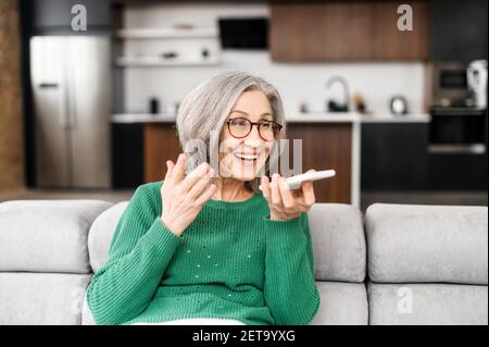 Reife Geschäftsfrau Großmutter mit grauem Haar in grünem Pullover, sitzt auf der Couch im Wohnzimmer und hält Mobiltelefon, spricht mit Lautsprecher an, gestikiert, Sprachnachricht aufzeichnen Stockfoto