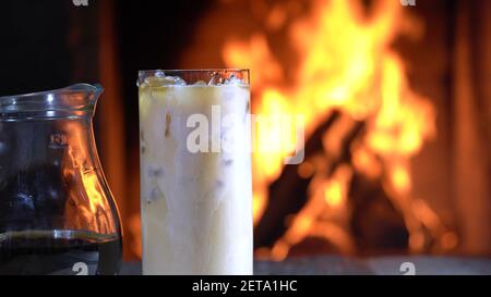 Milch gießt in ein Glas Kaffee mit Eis vor dem brennenden Kamin in einem Landhaus. Stockfoto