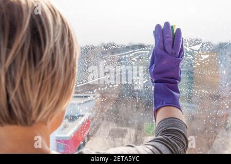 Eine unerkennbare junge Frau in blauen Handschuhen wäscht ein staubiges Fenster. Stockfoto