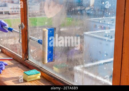 Eine unerkennbare junge Frau in blauen Handschuhen wäscht den Balkon Fenster von außen mit einem speziellen Langstreichschaber Stockfoto