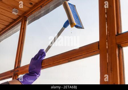 Eine unerkennbare junge Frau in blauen Handschuhen wäscht den Balkon Fenster von außen mit einem speziellen Langstreichschaber Stockfoto