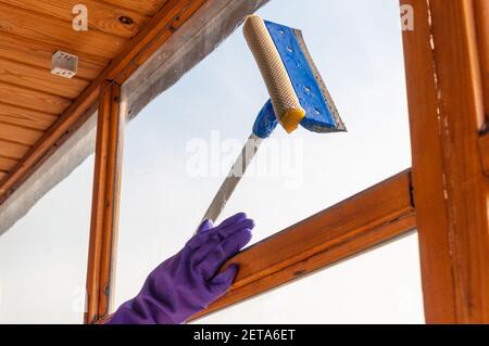 Eine unerkennbare junge Frau in blauen Handschuhen wäscht den Balkon Fenster von außen mit einem speziellen Langstreichschaber Stockfoto