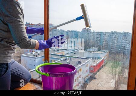 Eine unerkennbare junge Frau in blauen Handschuhen wäscht den Balkon Fenster von außen mit einem speziellen Langstreichschaber Stockfoto