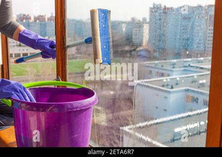 Eine unerkennbare junge Frau in blauen Handschuhen wäscht den Balkon Fenster von außen mit einem speziellen Langstreichschaber Stockfoto