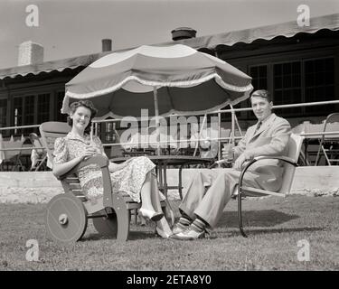 1940S PAAR MANN UND FRAU MANN FRAU SITZEN IM RASEN STÜHLE TRINKEN ALKOHOLFREIE GETRÄNKE UNTER EINEM SONNENSCHIRM LÄCHELND BLICK AUF CAMERA - F5707 HAR001 HARS ALTE MODE 1 STIL JUNG ERWACHSENE URLAUB FREUDE LIFESTYLE ZUFRIEDENHEIT FRAUEN VERHEIRATET EHEGATTEN EHEMÄNNER GESUNDHEIT KOPIEREN RAUM FREUNDSCHAFT GANZKÖRPERFRAUEN PERSONEN MÄNNER B&W PARTNER BLICKKONTAKT RESORT ZEIT AUS ANZUG UND KRAWATTE GLÜCK FRÖHLICH STYLES REISE KURZURLAUB ERHOLUNG RELAXD COUNTRY CLUB SODAS URLAUB LÄCHELT FRÖHLICHE STILVOLLE ZWEIFARBIGE MODEN PRIVILEG ENTSPANNUNG ZWEISAMKEIT FERIEN EHEFRAUEN JUNG ERWACHSENE MANN JUNGE ERWACHSENE FRAU Stockfoto