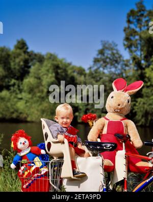 1980S BLONDE MÄDCHEN MIT RAGGEDY ANN PUPPE UND GEFÜLLT KANINCHEN SPIELZEUG SITZEN IN HINTEREN BABYSITZ AUF FAHRRAD SUCHEN BEI DER KAMERA - KB19941 GRD001 HARS KOPIEREN RAUM HALBE LÄNGE MÄNNCHEN FAHRRÄDER TRANSPORT FAHRRÄDER SOMMER BLICKKONTAKT GLÜCK ABENTEUER UND BUNNY RÜCKSITZ RAGGEDY ANN JUVENILES RAGGEDY SAISON ANN BABY MÄDCHEN BABYSITZ KAUKASISCHE ETHNIE ALTMODISCH Stockfoto