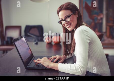 Glückliche junge intelligente kaukasische Frau toothy Lächeln zu Hause im Büro tippen auf Laptop, Blick auf die Kamera Stockfoto
