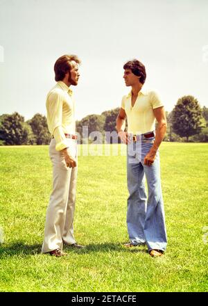 1970S ZWEI JUNGE MÄNNER MIT GLOCKENHOSE STEHEN AUF GRASSY HÜGEL REDEN ZUSAMMEN - KS12699 HAR001 HARS MÄNNCHEN GESCHWISTER STYLES GLOCKE UNTEN AUF GESCHWISTERHOSEN KONZEPTIONELLE GRASIGE STILVOLLE MODE ZWEISAMKEIT JUNGER ERWACHSENER MANN KAUKASISCHE ETHNIE HAR001 ALTMODISCH Stockfoto