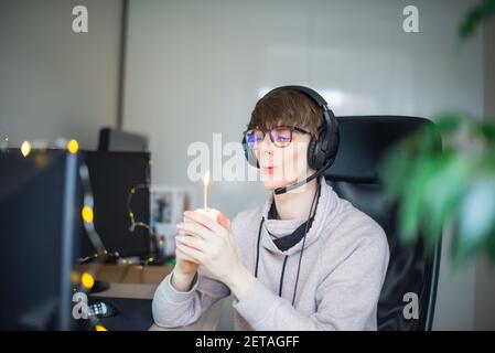 Junger Mann bläst Kerze auf dem festlichen Cupcake auf virtuelle Geburtstagsparty während des Lockdown. Generation Z Mann feiert seinen Geburtstag allein auf Stockfoto