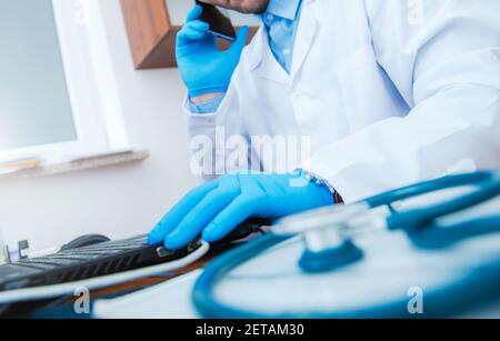 Kaukasischer Arzt Krankenhaus Büro Schreibtisch mit Stethoskop. Medizinischer Fachmann, der auf seinem Telefon spricht und Forschungsdaten auf seinem Laptop Com überprüft Stockfoto