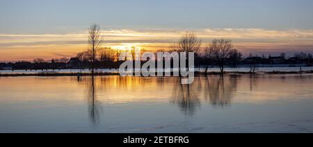 Silhouetten von kargen Bäumen im Winter bei farbenprächtiger Sonnenuntergangsstimmung Das Wasser des Flusses IJssel, wo die Auen haben Gefüllt Stockfoto