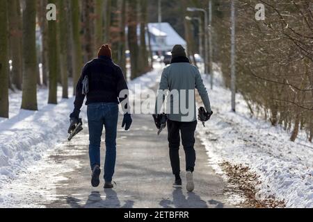 ZUTPHEN, NIEDERLANDE - 14. Feb 2021: Ende der Eislaufsaison mit zwei Männern von hinten gesehen, die mit ihren Schlittschuhen in Händen auf einer schneefreien Fahrbahn laufen Stockfoto