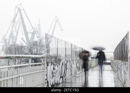 DANZIG, POLEN - Nov 13, 2018: Ein Paar mit Schirmen zu Fuß auf einer Brücke in Danzig Shipyard Station, Werftkrane im Hintergrund, eine horizontale Stockfoto