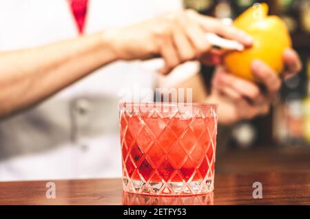 Nahaufnahme der Barkeeper-Hände, die den negroni-Cocktail mit Grapefruit zubereiten. Er setzt etwas Essenz aus Grapefruithaut in das Cocktailglas auf der Theke Stockfoto