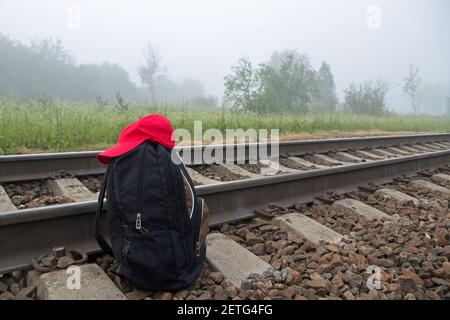 Schwarzer geschlossener Rucksack mit roter Baseballmütze auf der Oberseite Es steht auf dem braunen Granitschindel neben dem Schienen und Schwellen der Bahn gegen eine Stockfoto