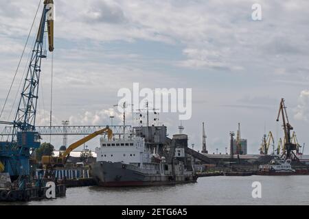 Kohleverladung oder -Entladung eines grauen Schüttgutfragers mit weißem Deck mit einem Bagger im Hafen, nahe gelegenen Ladeplattformen und Hafenkranen ca. Stockfoto