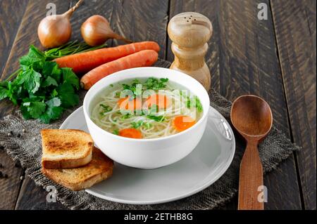 Frische Bouillon in einer weißen Schüssel mit fein gehackten Kräutern mit Petersilie und Knoblauch auf einem Holztisch. Heiße Suppe aus Hühnerfleisch, Karotten, Zwiebeln, Stockfoto