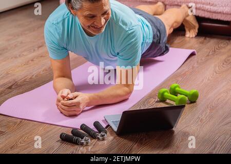 Der 50-jährige Mann führt Übungen durch, während er zu Hause auf dem Teppich liegt und den Computer anschaut. Während einer Pandemie trainiert eine Person in einer Wohnung über den Stockfoto