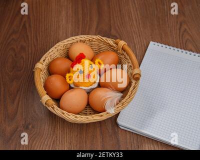 Gelbes Spielzeug-Huhn zwischen braunen Eiern im Weidenkorb neben dem Notizbuch auf brauner Oberfläche. Stockfoto