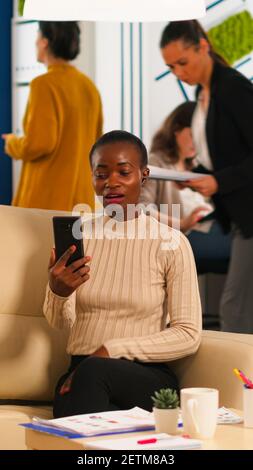 Müde schwarze Frau im Gespräch mit Kollegen mit Videoanruf halten Smartphone mit Wiraless-Headset sitzen auf der Couch im Büro. Vielfältiges Team plant neues Finanzprojekt in modernen Unternehmen Stockfoto
