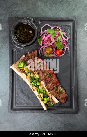 Rindersteak mit Zuckerknochen, serviert mit Sauce und Salat aus Zwiebeln, Pomiloren und Paprika. Dunkler Hintergrund Stockfoto