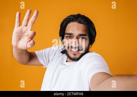 Lächelnder indischer Mann nimmt ein Selfie isoliert über gelben Hintergrund, lässige Kleidung, ok Geste Stockfoto