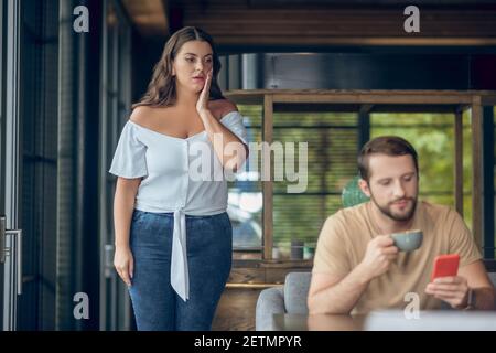 Überraschte Frau in der Nähe von Tisch und Mann mit Smartphone Stockfoto