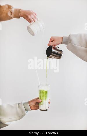 Weibliche Hände Gießen japanischen Matcha Tee und Milch aus Krug in Glas für die Herstellung von grünen Matcha Latte zum Frühstück über helle Wand Hintergrund. Kaffee Getränke Getränke und japanische Küche Stockfoto