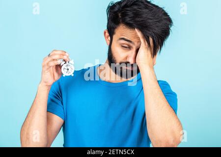 Emotionales Porträt eines gutaussehenden, verschlafenen Hindu-Mannes mit kleinem Wecker in einem blauen T-Shirt auf hellblauem Hintergrund Stockfoto