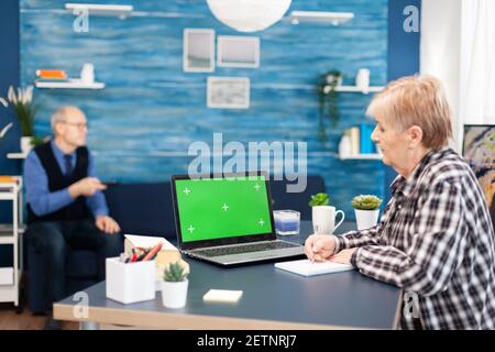 Ältere Frau, die sich Notizen zu einem Notebook mit einem tragbaren Computer mit Kopierplatz gemacht hat. Ältere Frau, die an einem Laptop mit grünem Bildschirm arbeitet, und Ehemann, der die fernbedienung hält. Stockfoto