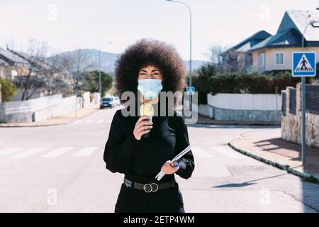 Fernsehjournalistin mit Afro-Haaren, die eine chirurgische Maske trägt, um sich vor einem Virus zu schützen, und berichtet über eine Geschichte auf der Straße Stockfoto