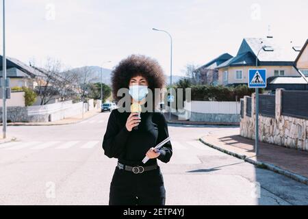 Fernsehjournalistin mit Afro-Haaren, die eine chirurgische Maske trägt, um sich vor einem Virus zu schützen, und berichtet über eine Geschichte auf der Straße Stockfoto