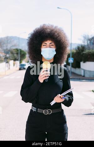 Fernsehjournalistin mit Afro-Haaren, die eine chirurgische Maske trägt, um sich vor einem Virus zu schützen, und berichtet über eine Geschichte auf der Straße Stockfoto