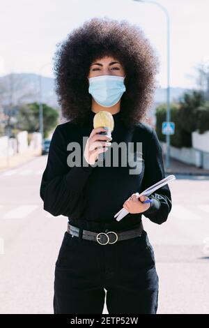 Fernsehjournalistin mit Afro-Haaren, die eine chirurgische Maske trägt, um sich vor einem Virus zu schützen, und berichtet über eine Geschichte auf der Straße Stockfoto