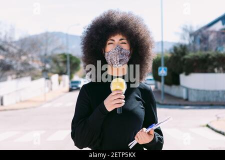 Fernsehjournalistin mit Afro-Haaren, die eine Leopardenhaut-Maske trägt, um sich vor einem Virus zu schützen und über eine Geschichte auf der Straße berichtet Stockfoto