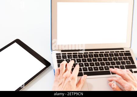 Hände einer Frau, die auf einem Laptop neben einem Tablet tippt, mit weißen Bildschirmen zum Einfügen von Designs oder Nachrichten, alles auf weißem Hintergrund Stockfoto